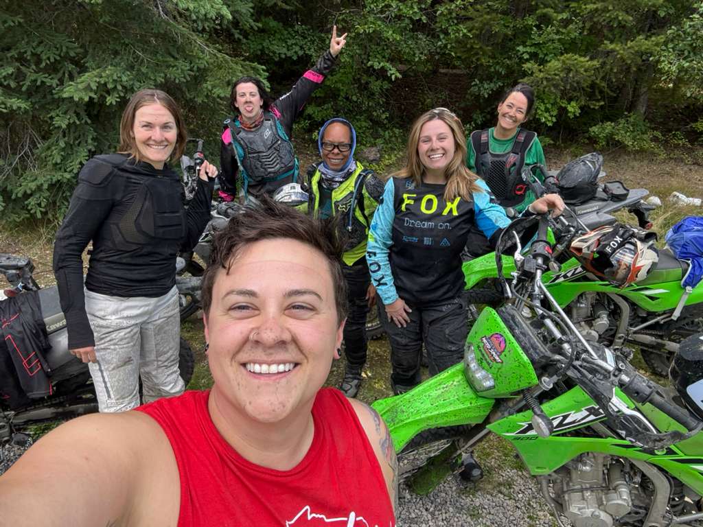 Group of six smiling women motorcyclists posing for a selfie outdoors with green dirt bikes and forest background.
