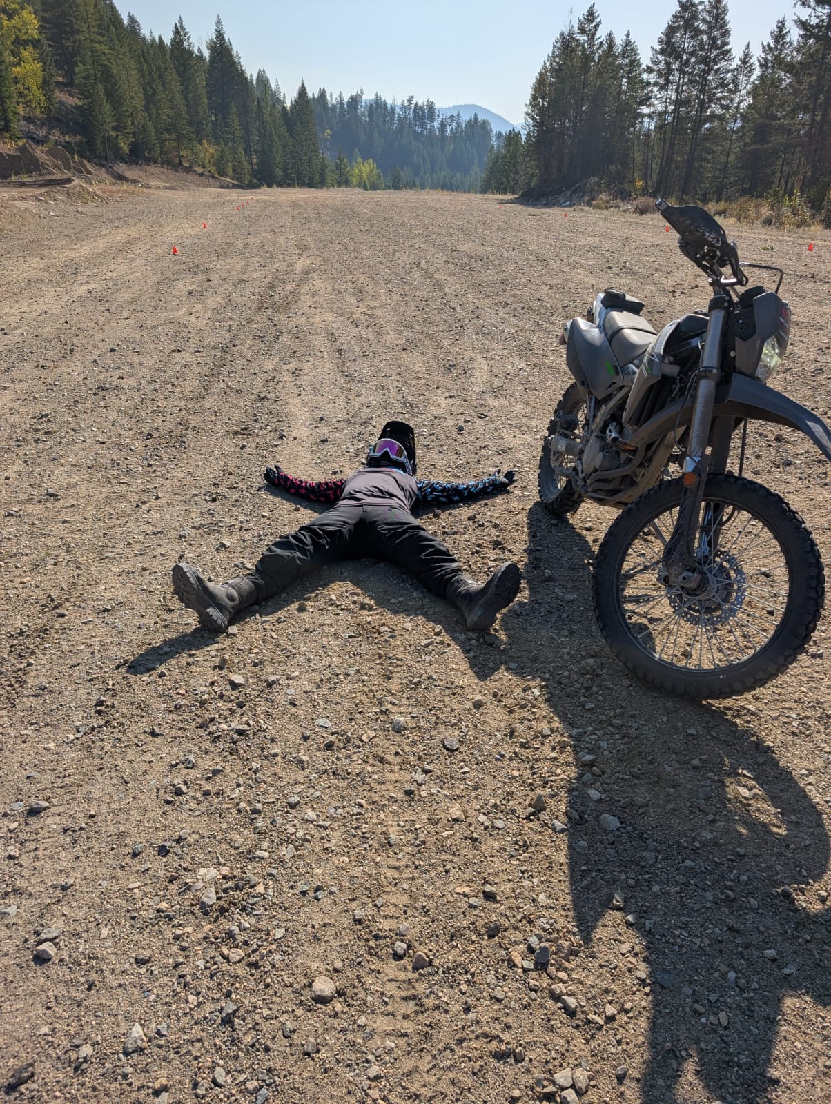 Woman wearing motocross gear lying on dirt road beside a parked dirt bike in a forested area.