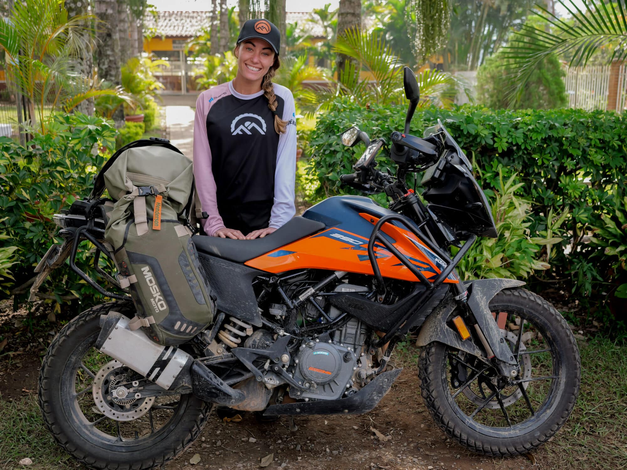 Young woman with a braided ponytail wearing a cap and black and pink shirt standing next to an orange and black dirt bike with a green Mosko Moto bag attached, in a lush garden setting.