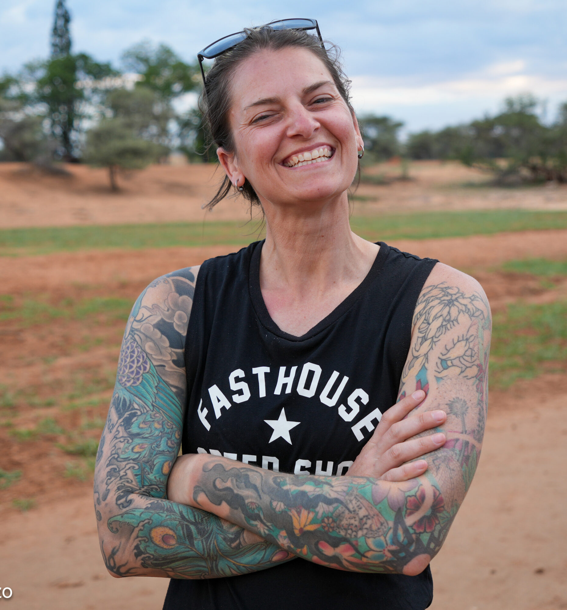 Smiling woman with colorful arm tattoos wearing a black sleeveless shirt and sunglasses on her head standing outdoors on a dirt path with trees in the background.
