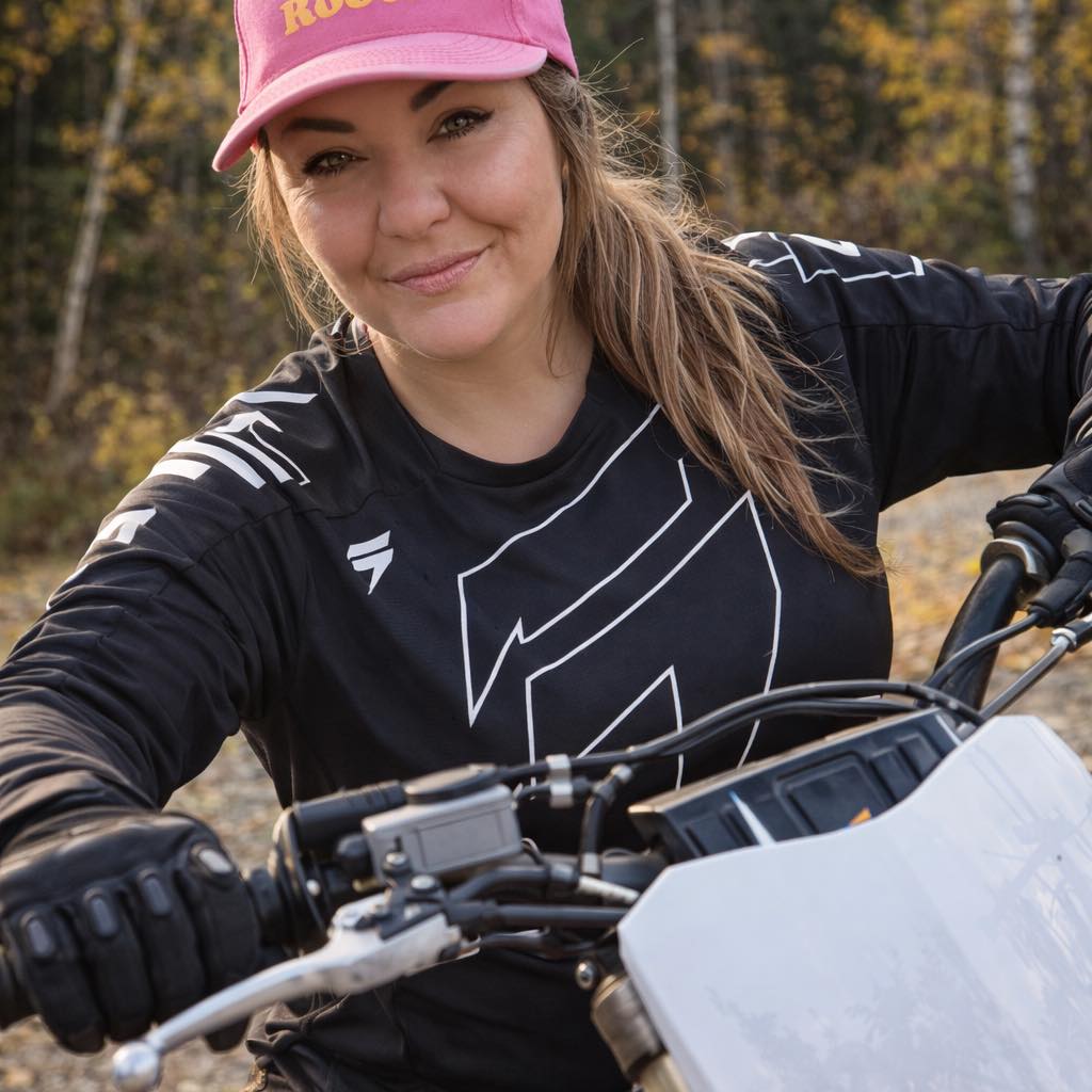 Woman wearing a pink cap and black motocross gear riding a dirt bike outdoors in autumn.