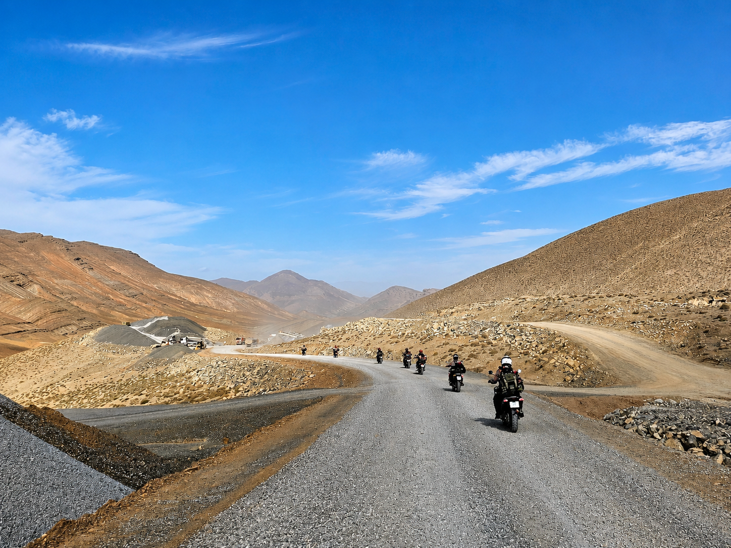 Motorcyclists riding along a winding gravel road through a mountainous landscape under a clear blue sky, showcasing the beauty of adventure travel in nature.