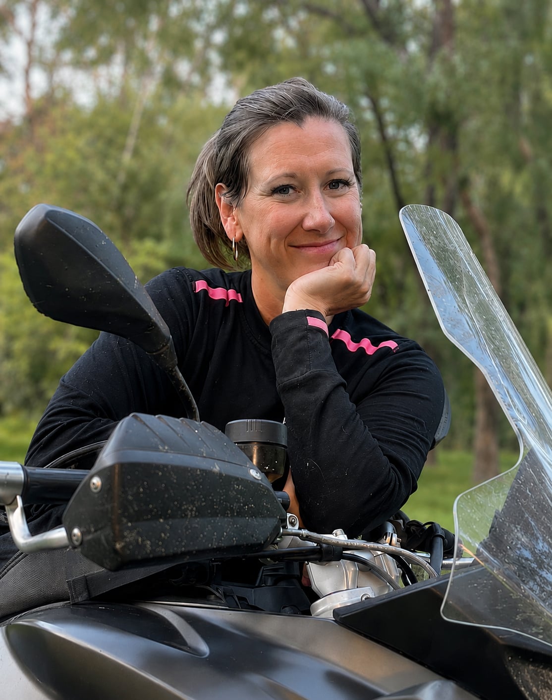 Smiling woman resting her chin on her hand while sitting on a motorcycle, surrounded by greenery.