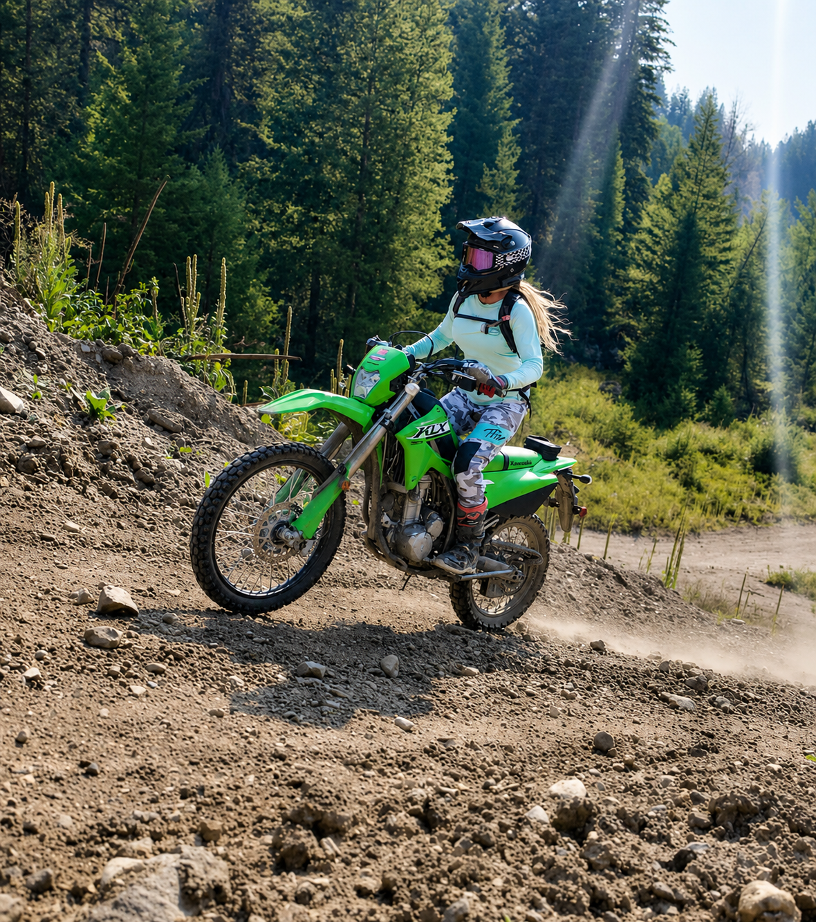 Woman riding green Kawasaki KLX dirt bike on rough dirt trail in forested mountain area with helmet and protective gear.