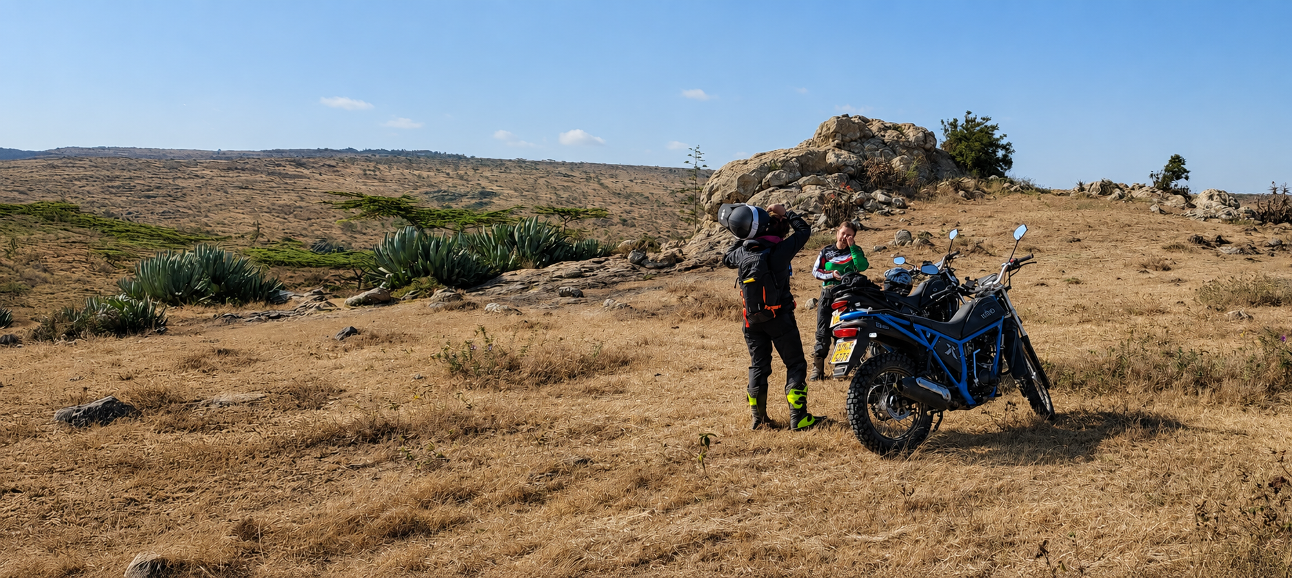 Two motorcyclists preparing for a ride in a scenic, dry landscape with rocky formations and sparse vegetation. One rider is adjusting their helmet while the other stands beside a motorcycle, showcasing outdoor adventure and motorcycle culture.