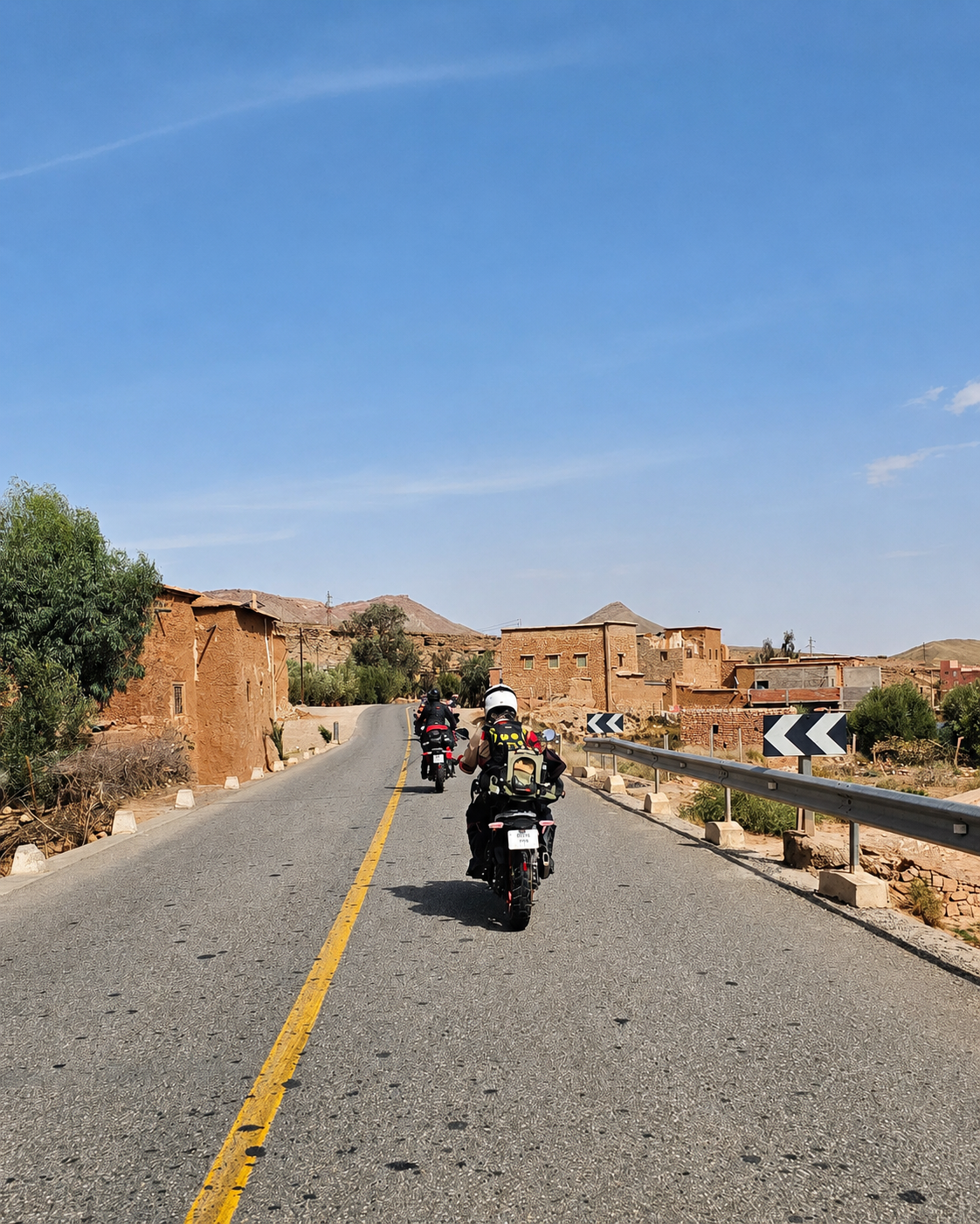 Motorcyclists riding on a paved road through a desert village with adobe buildings under a clear blue sky.